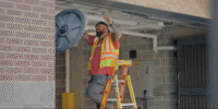 Construction worker in a neon safety vest on a yellow ladder, working on exposed ceiling utilities in a basement utility room