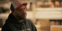 Portrait of a man wearing a red-and-gray trucker cap and black hoodie, looking contemplative in a softly lit indoor setting.