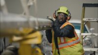 Worker in a high-visibility vest and hard hat leans on a large metal pipe in an industrial setting, wearing protective gear (glasses and ear protection).